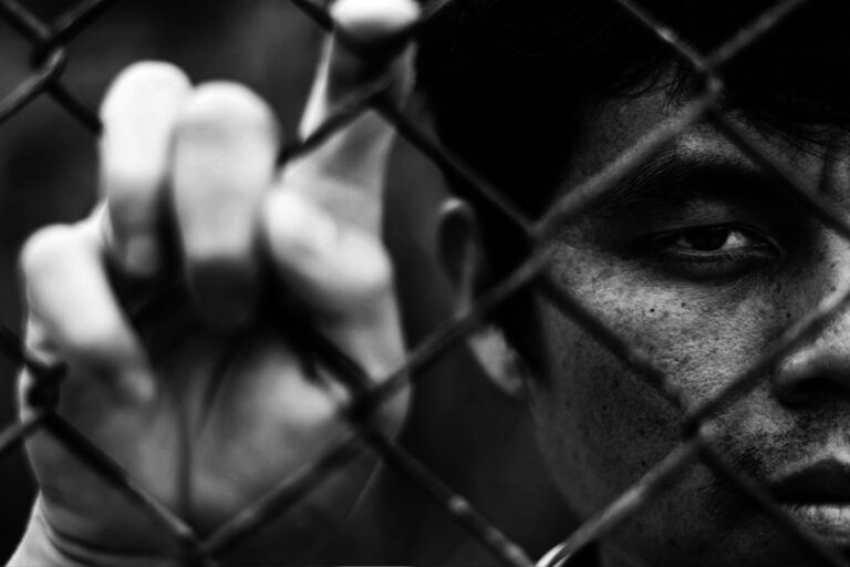 Depressed man standing behind a fence, hand grabs steel mesh cage,close up on face in white tone, abuse concept